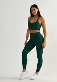 Ciemnozielony stanik sportowy i legginsy o wysokim stanie wykonane z elastycznego materiału, w zestawieniu z białymi butami sportowymi. Gładka faktura, minimalistyczny design.