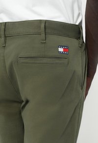 Zaļas kokvilnas bikses ar aizmugurējo kabatu, kurā ir "TOMMY JEANS" logo plāksne sarkanā, baltā un zilā krāsā. Vienkārša dizaina, gluda tekstūra.