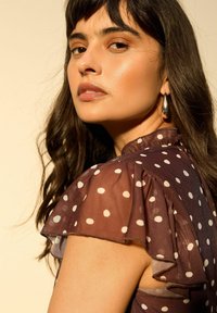 Donna con capelli lunghi e scuri che indossa orecchini a cerchio d'argento e una maglia marrone trasparente a pois, che guarda oltre la spalla contro uno sfondo neutro.