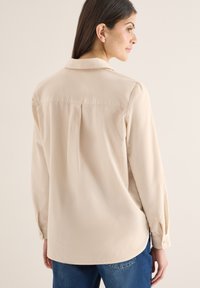 Femme aux cheveux bruns portant une chemise beige à manches longues et un jean bleu, vue de dos et de profil, sur un fond clair uni.