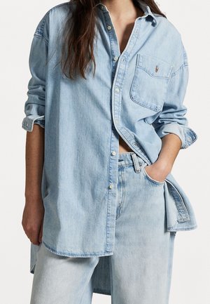 Osoba ubrana w dużą jasno-niebieską dżinsową koszulę z podwiniętymi rękawami oraz jasno-niebieskie, wysokie spodnie dżinsowe, z ręką w kieszeni, stojąca na białym tle.