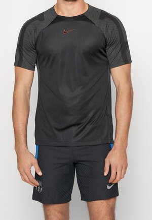 Homme portant un maillot de sport noir Nike avec des manches gris foncé à motifs et un short noir avec des bandes bleues et des logos d'équipe.