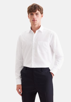 Jeune homme portant une chemise blanche à manches longues boutonnée et un pantalon noir, debout avec une main dans la poche, fond neutre.