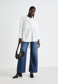 Donna alta che indossa una camicia bianca a righe oversized, jeans a gamba larga blu, stivaletti neri alla caviglia, mentre tiene una piccola borsa a tracolla marrone scuro.