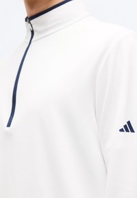 Λευκό πουλόβερ με μακριά μανίκια, μπροστινό φερμουάρ τέταρτου μήκους σε ναυτικό μπλε και λογότυπο σε ναυτικό μπλε στο αριστερό μανίκι, φορεμένο από άτομο που δείχνει τον λαιμό και το άνω μέρος του κορμού.