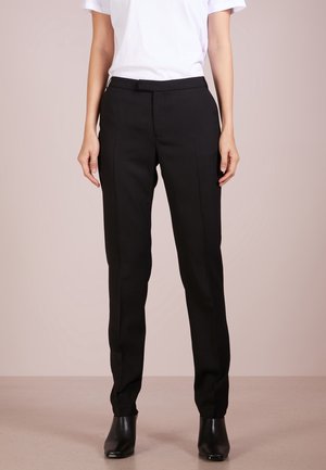 Osoba nosząca dopasowane czarne spodnie, czarne botki za kostkę oraz gładką białą bluzkę z krótkim rękawem, stojąca na neutralnym tle.