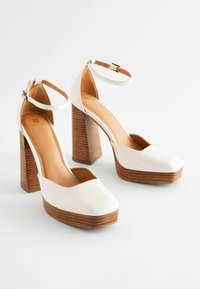 Chaussures à talons hauts blanches avec plateforme et talon en bois. Elles sont dotées d'une bride à la cheville et d'une texture lisse et brillante. Design à bout pointu.