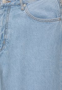 Blugi de denim bleu deschis cu o textură netedă, având un buzunar frontal și cusături contrastante de culoare maro de-a lungul cusăturilor și taliei.