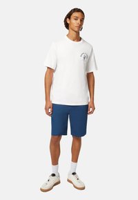 Hvid bomulds t-shirt med korte ærmer og et lille blåt logo, parret med blå shorts, hvide sokker og beige sneakers.