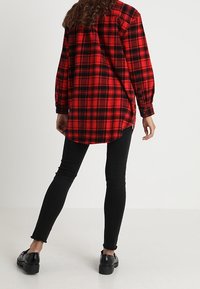 Camicia di flanella a quadri rossi e neri con maniche lunghe, orlo arrotondato, chiusura frontale a bottoni, abbinata a jeans skinny neri e scarpe nere.