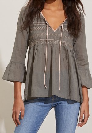 Femme portant un chemisier gris à manches longues avec une broderie rose et des liens, associé à un jean bleu clair, debout devant un fond uni.