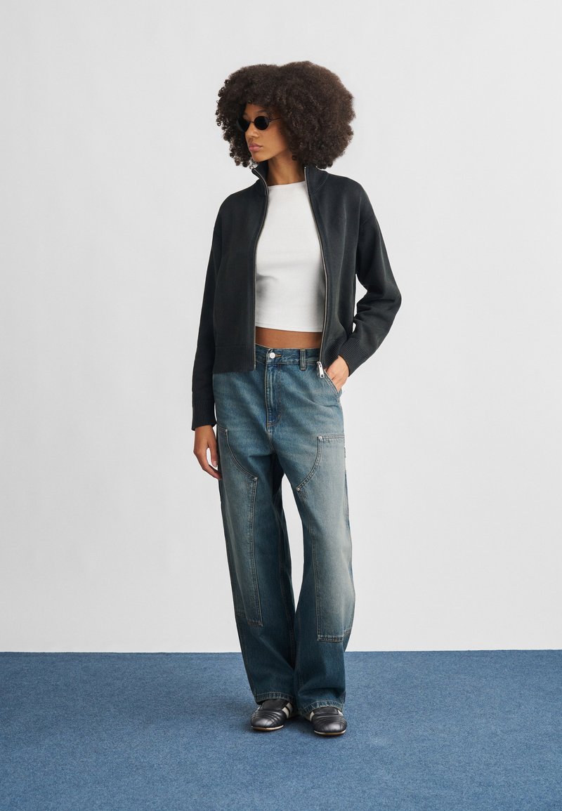 Sort lynlåsjakke, hvid crop top, lyseblå løstsiddende jeans. Model iført sorte solbriller, sølvfarvede sko med tre striber. Simpelt baggrund.