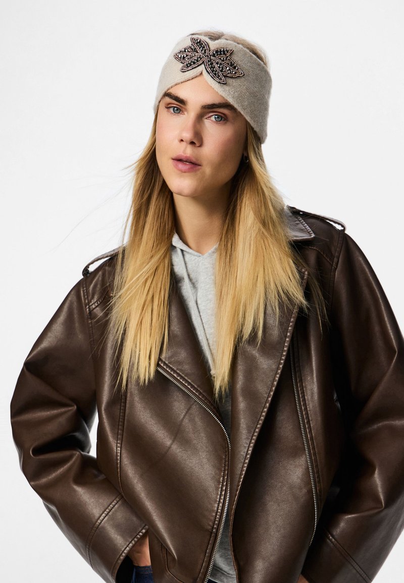 Beige kudumine peapael, millel on keerutatud disain, ning must ja hõbedane tikandiga lipsu aksessuaar. Kantud pruunist nahkjakist.