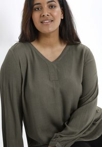 Olivi vihreä pitkähihaisen puseron V-pääntie ja hienovarainen tekstuuri, jossa pieni lovi yksityiskohta rinnassa. Liu'uttava kangas rento istuvuus.