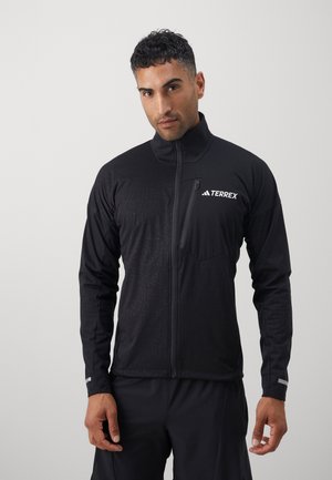 Man wearing black zip-up jacket with Terrex logo, standing against plain light background, facing forward with hands relaxed.
