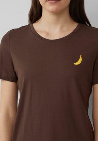 Bruine katoenen t-shirt met korte mouwen en een ronde halslijn. Bevat een kleine gele banaanborduring op het linkerborstgebied.