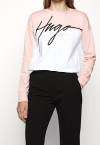 Vrouw in een roze en witte lange mouwen sweatshirt met zwarte scripttekst "Hugo" in combinatie met zwarte broek, met één hand in haar zak.