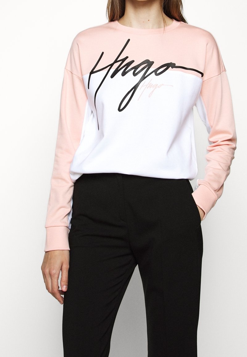 Vrouw in een roze en witte lange mouwen sweatshirt met zwarte scripttekst "Hugo" in combinatie met zwarte broek, met één hand in haar zak.