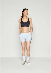 Jauna moteris stovi su rankomis kišenėse, vilkinti juodą sportinį liemenėlę, šviesiai mėlynas šortus, baltas kojines ir sportinius batelius prieš vienodą foną.