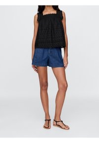 Kvinna klädd i ärmlös svart spets-top, blå denimshorts med knappar och svarta platta sandaler mot en ljus bakgrund.