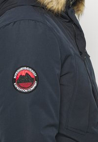 Granatowa kurtka z kapturem podszytym sztucznym futrem. Na górnej części rękawa znajduje się wyszywana okrągła naszywka w kolorach czerwonym i czarnym, przedstawiająca motyw górski.