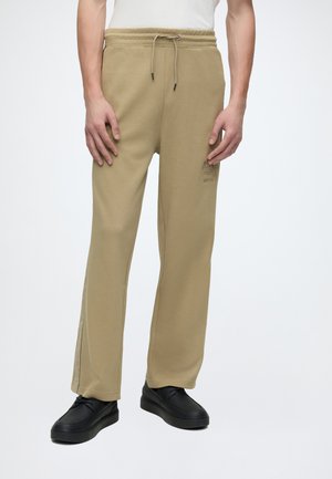 Uomo che indossa pantaloni della tuta beige con coulisse con un logo ricamato sottile sulla coscia sinistra e scarpe nere stringate.