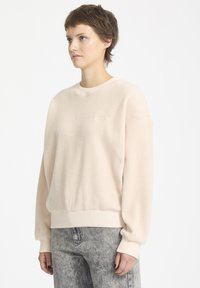 Jasno beżowy polarowy sweatshirt o teksturowanej powierzchni, z prążkowanymi mankietami i dołem, z subtelnym logo z przodu. W połączeniu z szarymi dżinsami.