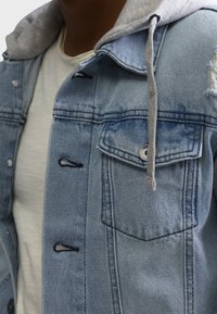 Chaqueta de mezclilla azul claro con capucha gris. Cuenta con bolsillos con botones, costuras visibles y un detalle desgastado en el hombro.