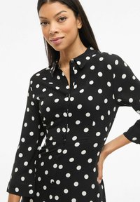 Černé šaty s puntíky a dlouhými rukávy, se zapínáním na knoflíky vpředu a klasickým límečkem. Hladký materiál s volným střihem.