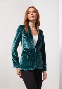 Teal velvet blazer with notched lapels, single button closure, and two front pockets. Worn over a white top and paired with black pants.