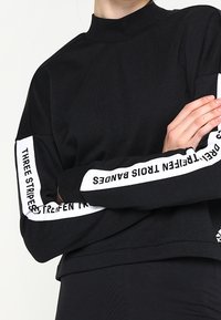 Sweatshirt noir à manches longues avec col montant, orné de rayures blanches sur les manches avec du texte, fabriqué dans un tissu lisse et texturé.
