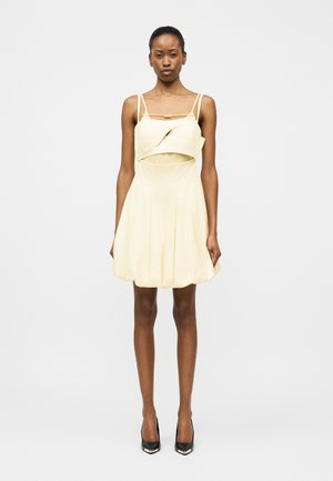 Mujer con un vestido mini sin mangas de color amarillo pálido con detalle de volantes en el frente y tacones altos negros, de pie contra un fondo blanco liso.