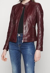 Giacca in pelle bordeaux con collo tondo, design aderente, cuciture visibili e due zip laterali. Abbinata a jeans blu slavati.