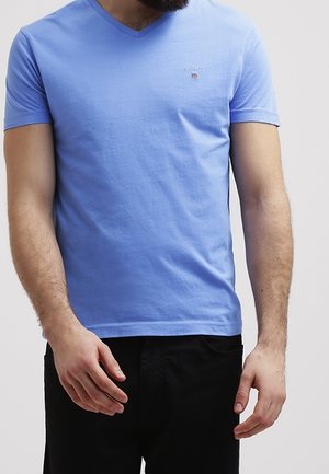 Homme portant un T-shirt bleu clair à manches courtes en V avec un petit emblème sur la poitrine, et un pantalon noir, debout devant un fond uni.