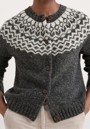 Dark gray wool cardigan with white geometric yoke pattern and brown buttons, worn with light beige corduroy pants, hands in pockets.