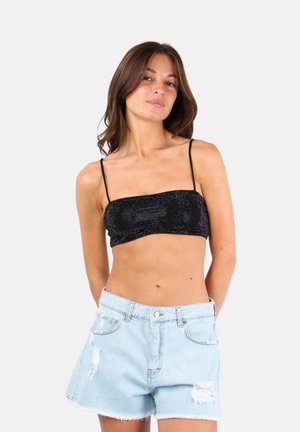 Giovane donna con capelli castani indossa un top corto nero con bretelle sottili e pantaloncini di jeans azzurri consumati su uno sfondo semplice.