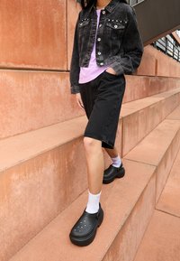 Schwarze Denim-Jacke mit silbernen Knöpfen, kombiniert mit einem helllila Hemd, schwarzen Shorts und schwarzen Clogs über weißen Socken auf Treppen.