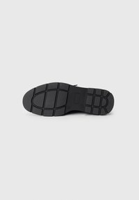 Black shoe sole with a textured, rubber surface featuring a grid pattern for traction. Round shape and flat heel design.