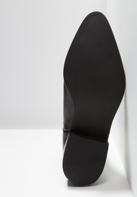 Semelle de chaussure de ville en cuir noir avec un talon rectangulaire texturé et un bout pointu, photographiée de dessous sur fond blanc.