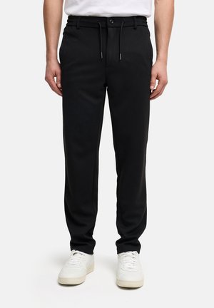 Uomo che indossa pantaloni neri con coulisse e sneakers bianche, in piedi su uno sfondo bianco uniforme.