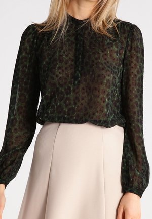 Woman wearing a sheer black long-sleeve blouse with a subtle leopard print and a high-neck collar paired with a beige skirt.