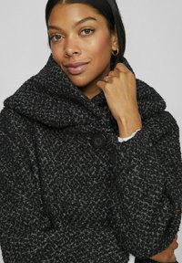 Manteau texturé noir et gris avec un grand col, doté d'une fermeture par un seul bouton et d'un design confortable. Tissu doux présenté en gros plan.