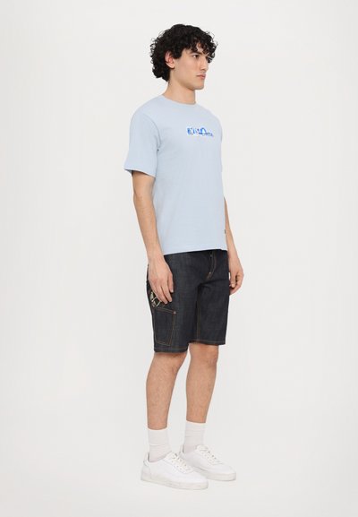 Light blue cotton t-shirt with graphic print, paired with dark denim shorts featuring a side pocket and white sneakers.