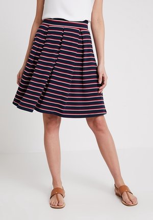 Mujer con falda azul marino con rayas horizontales blancas y rojas, camiseta blanca y sandalias marrones abiertas en los dedos, de pie frente a un fondo liso.