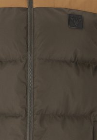 Brown and olive green puffer jacket featuring quilted sections, a central zipper, and a black logo patch on the left side.