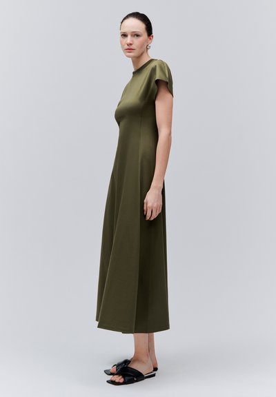 Femme aux cheveux foncés portant une robe midi vert olive à manches courtes et des sandales noires à enfiler, debout devant un fond uni gris clair.