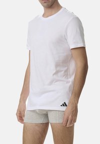 White short-sleeve t-shirt in cotton with a crew neck, featuring a small black Adidas logo on the lower left. Designed for casual wear.