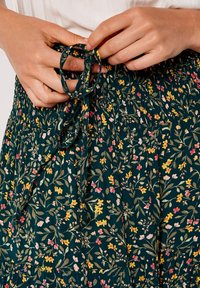 Floral skirt in dark green fabric with elastic waistband and tie detail. Features small yellow, pink, and white flowers, and leafy patterns.