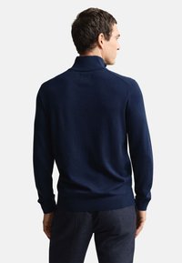 Marineblauwe gebreide pullover met een opstaande kraag en lange mouwen, met een gestructureerde afwerking en geribde manchetten en zoom, gezien vanaf de achterkant.