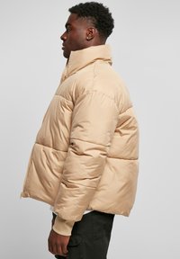 Beige buksejakke med høy krage, vertikal quiltet design og elastiske mansjetter. Har glidelås foran og en avslappet passform.